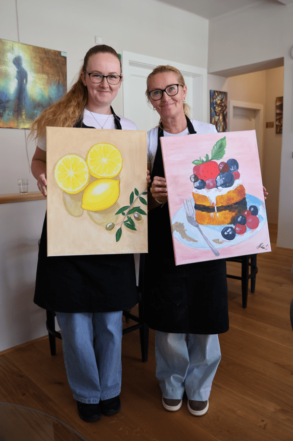 Dvě usměvavé ženy drží malované obrazy s mořem, hvězdou a plachetnicí v restauraci. Na kurzech M-Art.